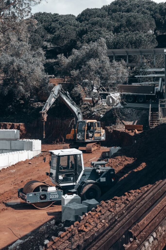 Excavators and vehicles operate on a forested construction site under a clear sky.